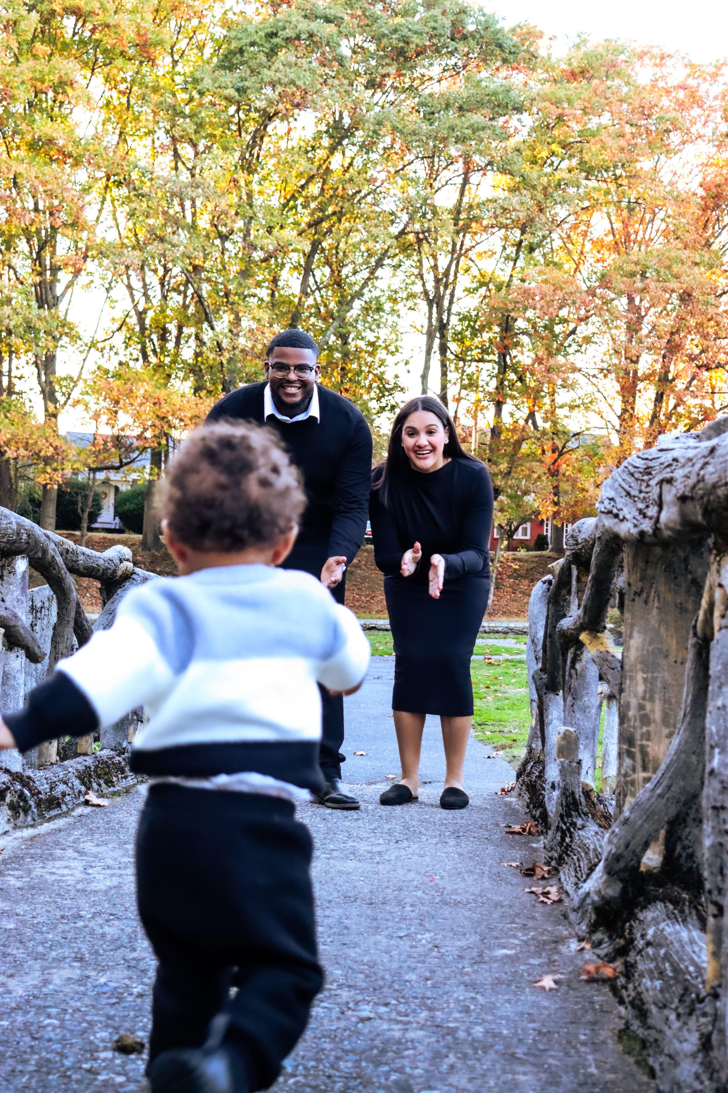 Two people smile with joy as a small child runs towards them on an autumn path lined with trees.