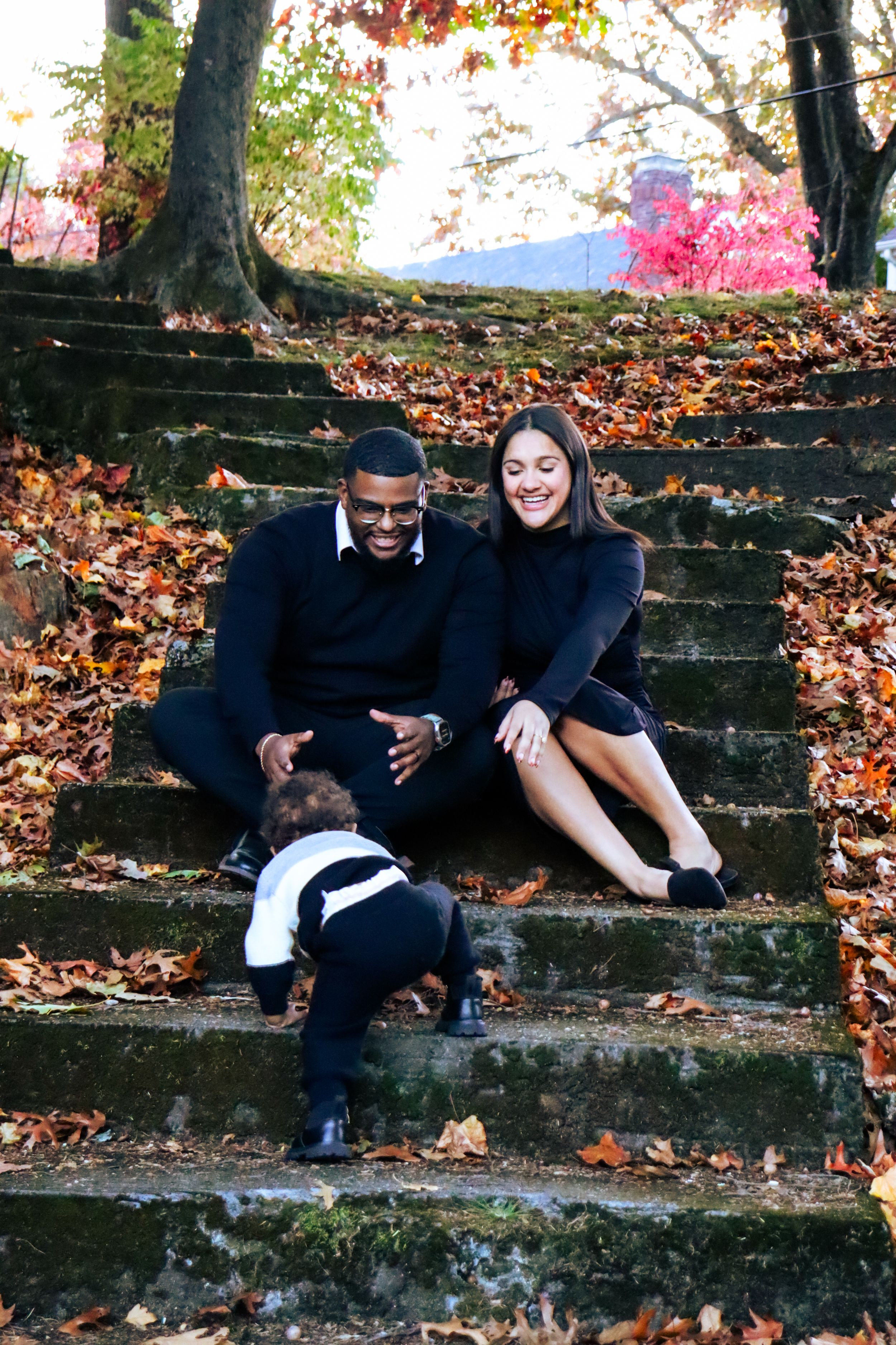 A couple sits on stone steps covered in fall leaves while a child plays below.