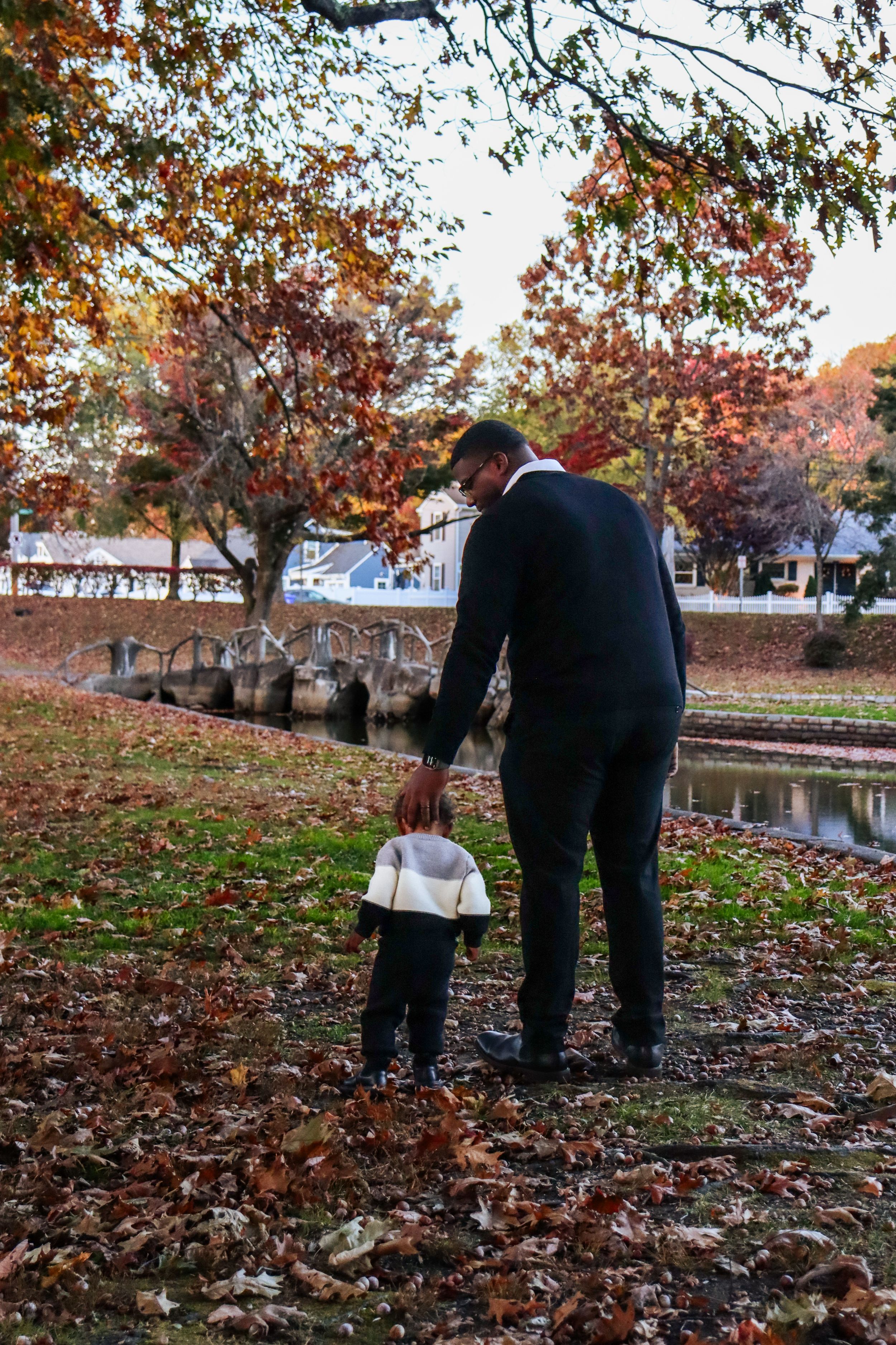 Adult and small child hold hands while walking through a park with autumn foliage.