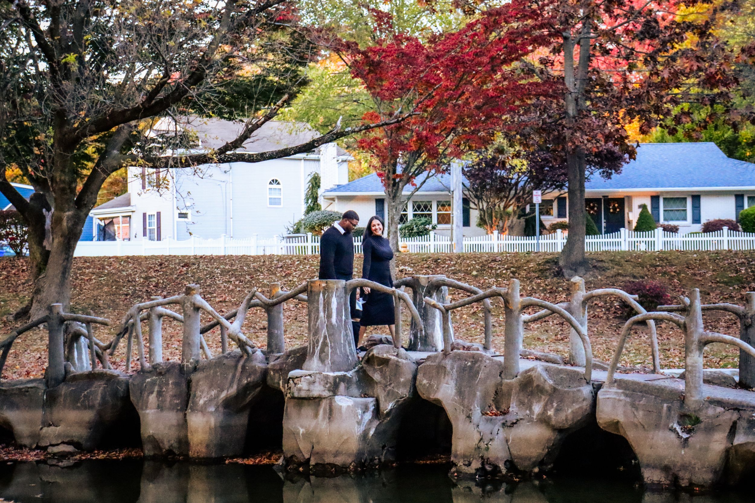 People stand on a rustic wooden bridge surrounded by vibrant fall colors and white houses.