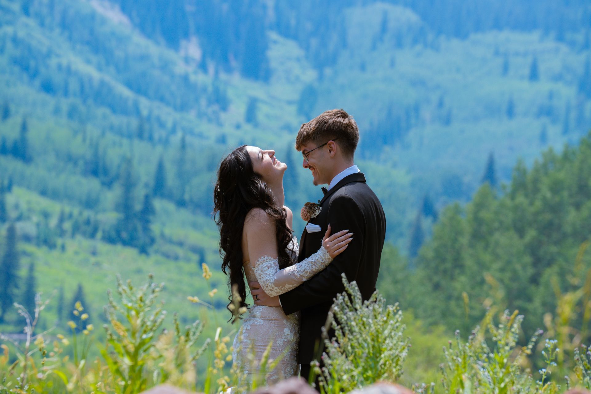 Bride and groom share a romantic embrace on a green mountain hillside during their ceremony.