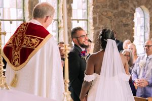 A wedding ceremony takes place in a rustic stone chapel with stained glass windows and white-veiled attendees.