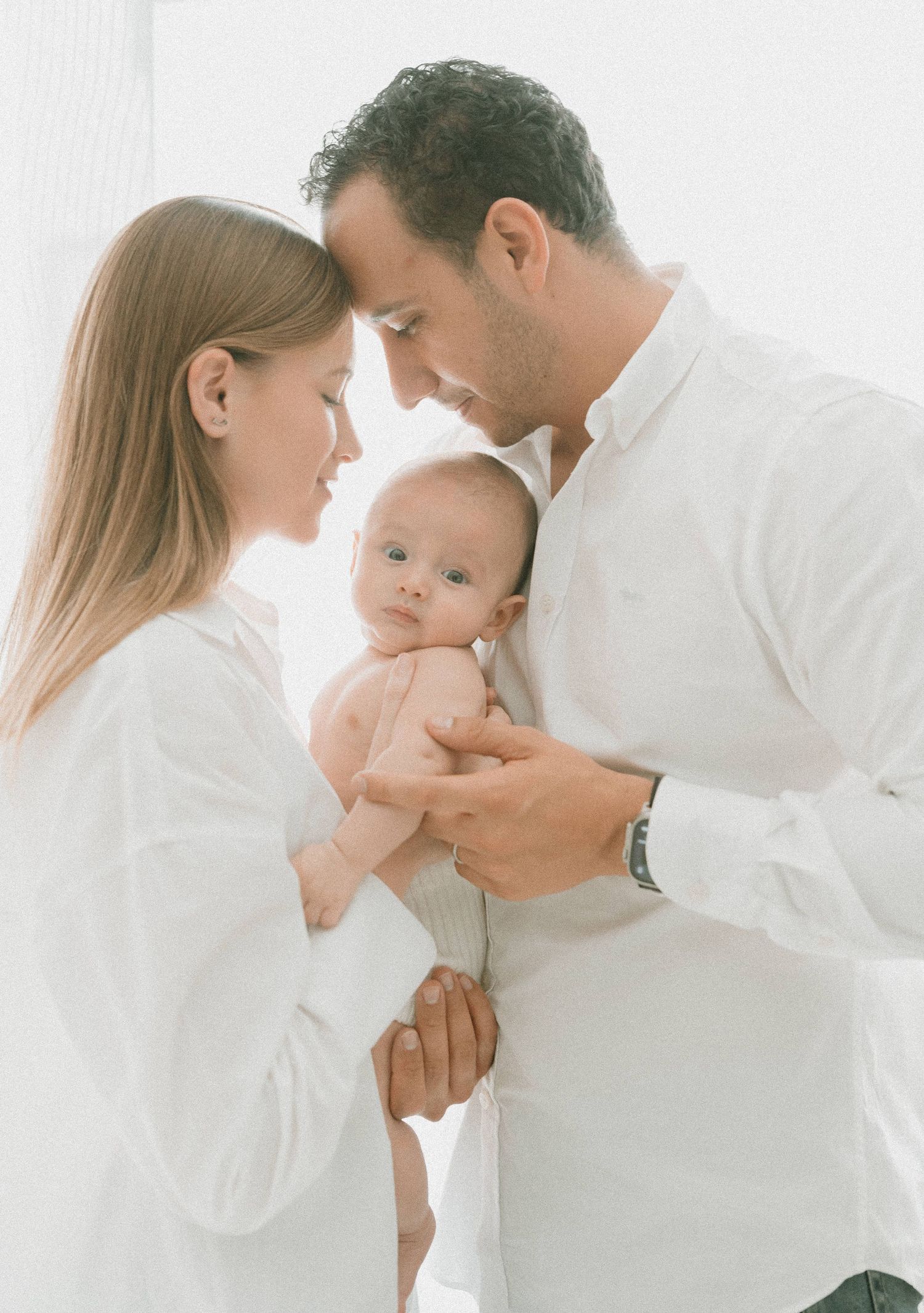 Intimate family portrait in white clothing with parents cradling baby between them.