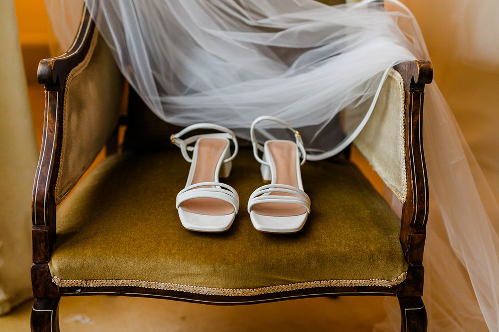 White wedding shoes on vintage velvet chair with flowing dress fabric.