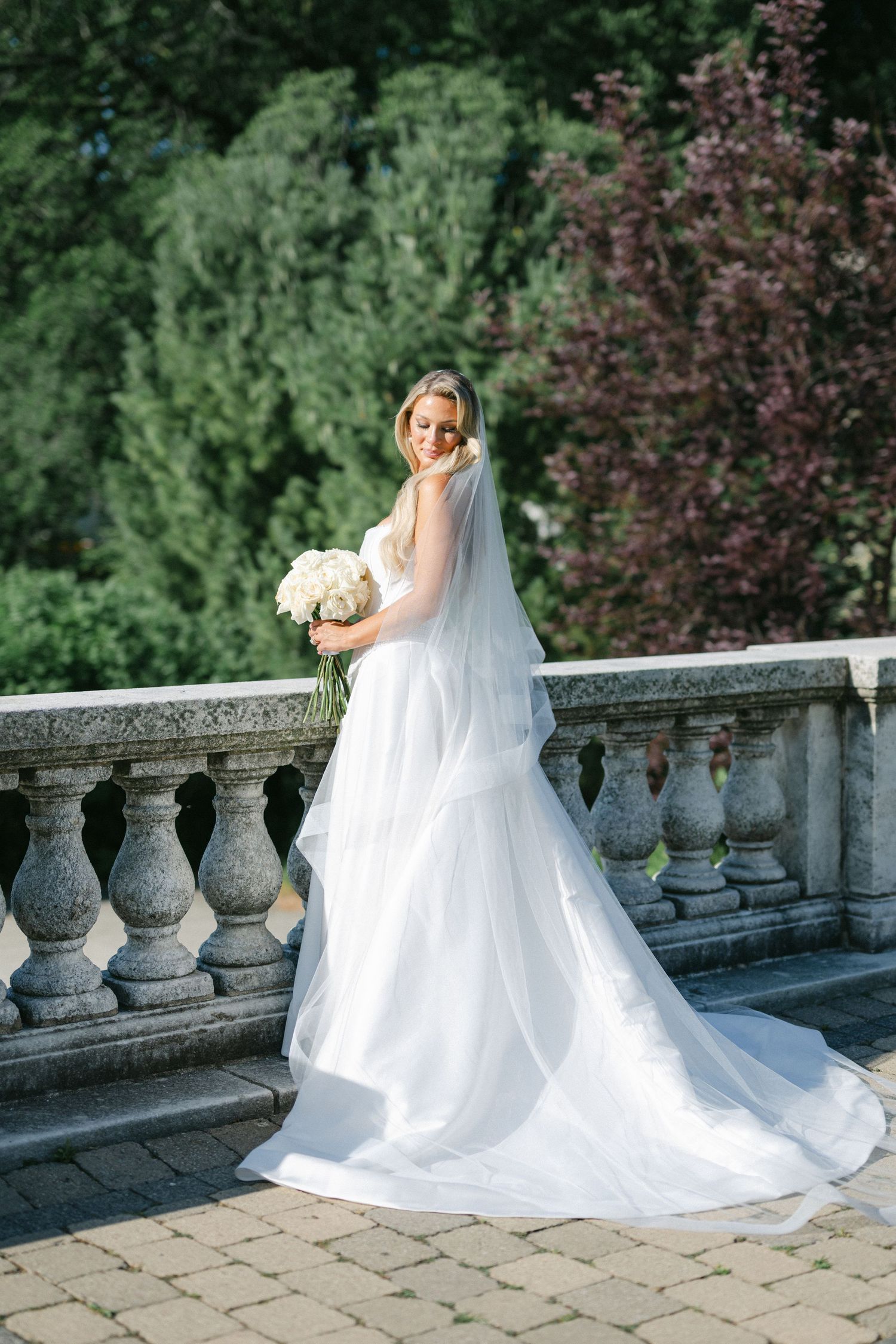 A flowing white wedding dress cascades down stone steps in a romantic garden setting, creating an elegant bridal portrait series.