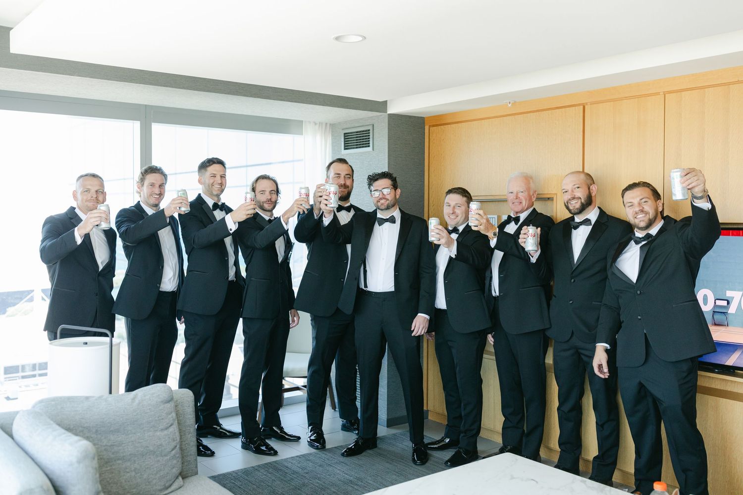 Groomsmen in black tuxedos and bowties stand in a row against a light colored wall at a formal wedding event.