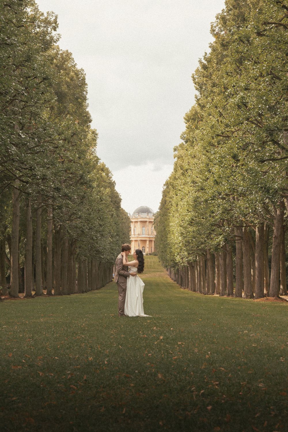 A couple embraces on a tree-lined path leading to a classical building at dusk.