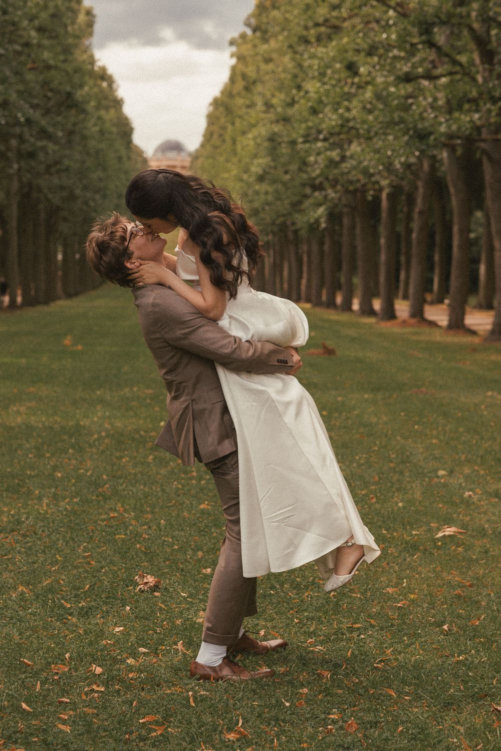 A romantic couple embraces and dances together on an autumn day in a tree-lined park with fallen leaves.