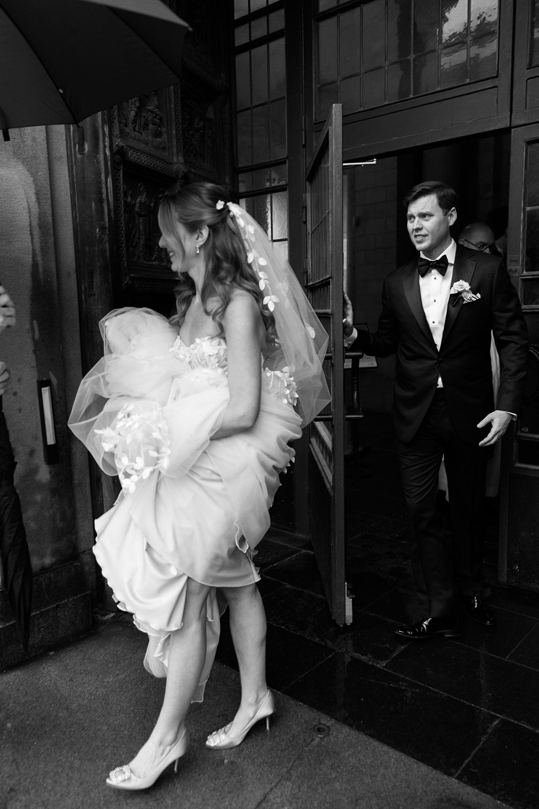 A bride in a white wedding dress lifts her gown while her groom watches from a doorway in this black and white photo.