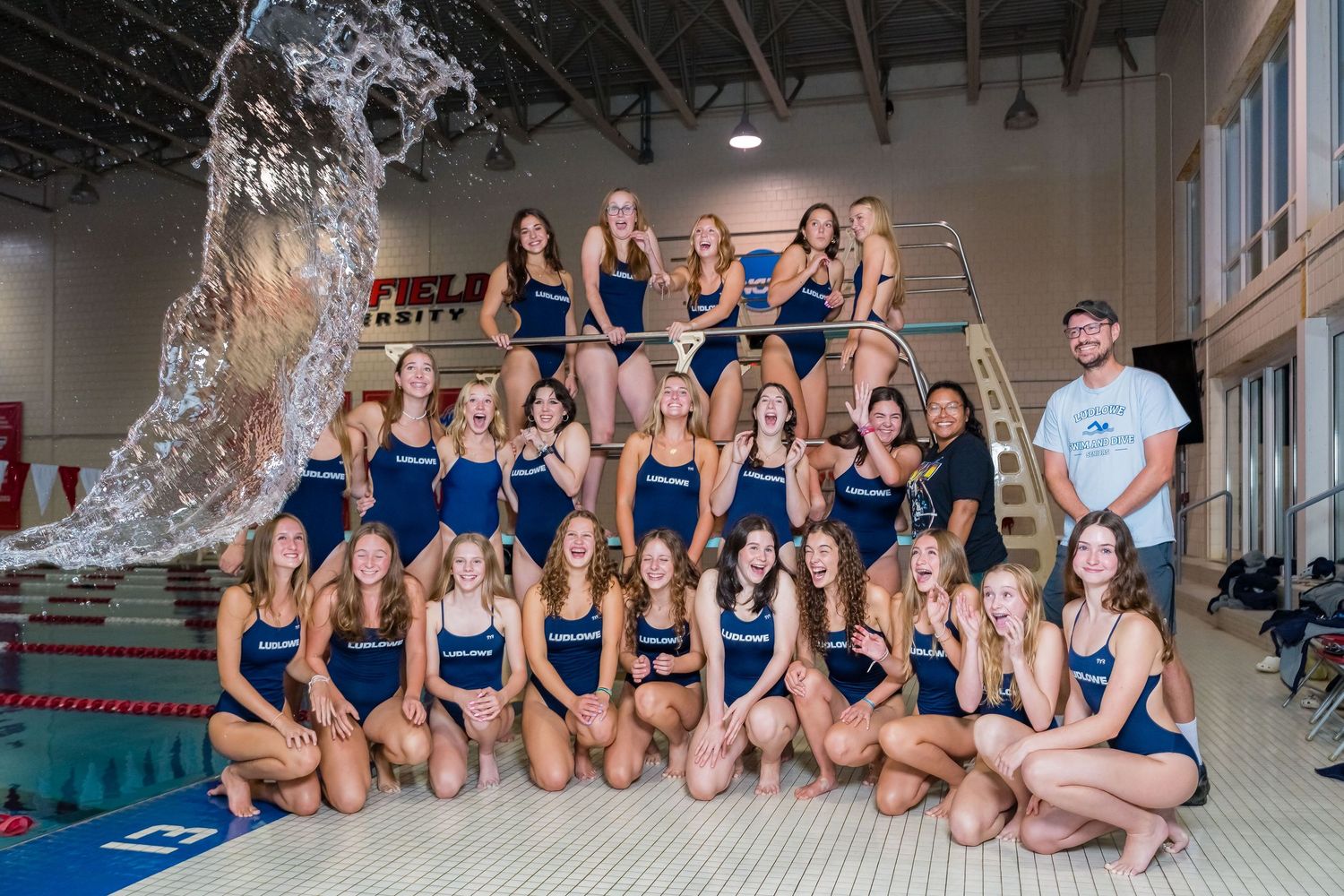A swim team poses together on pool deck stairs wearing matching navy blue swimsuits beside an indoor swimming pool.