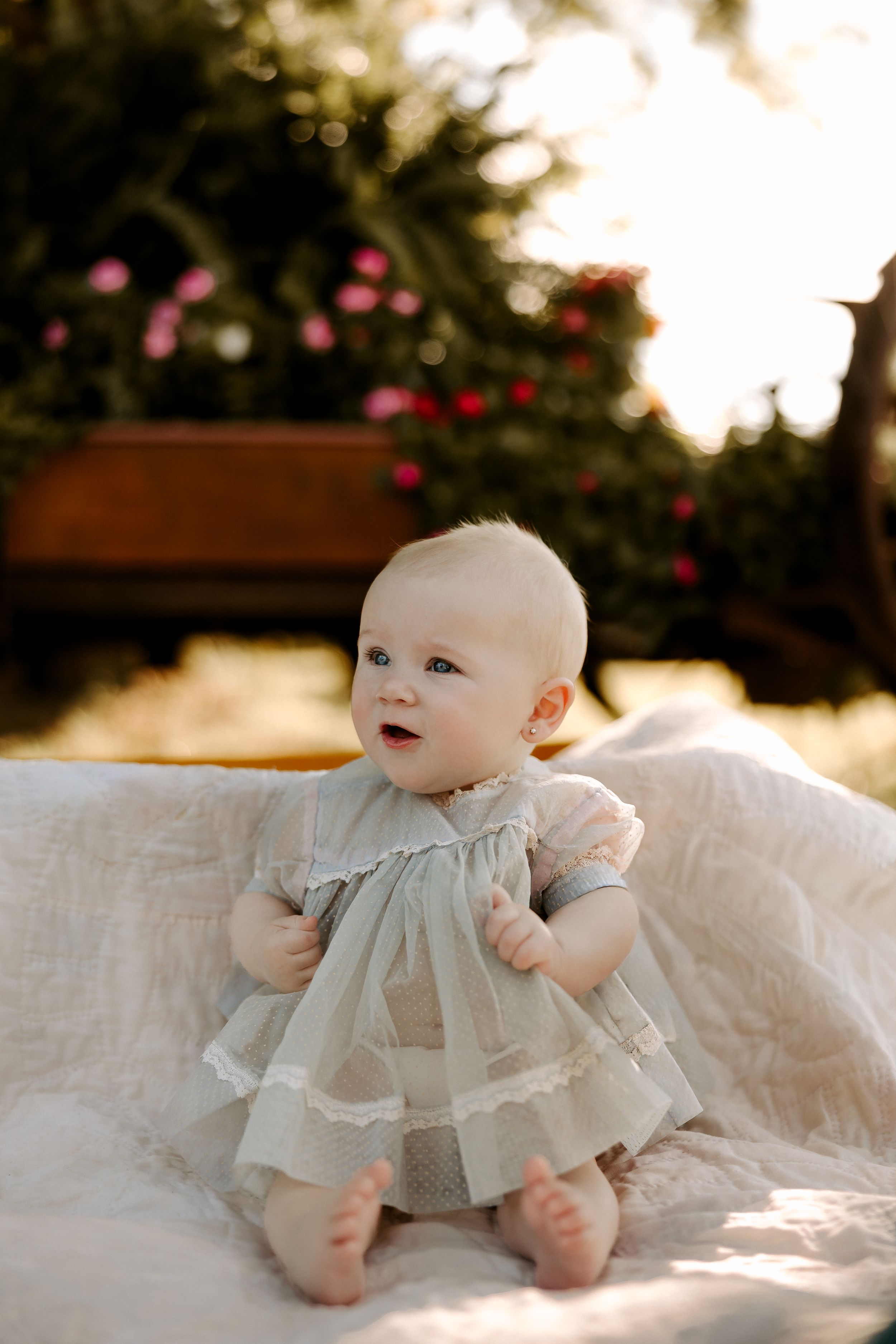 Baby in vintage-style dress lying on white blanket with trees in background.