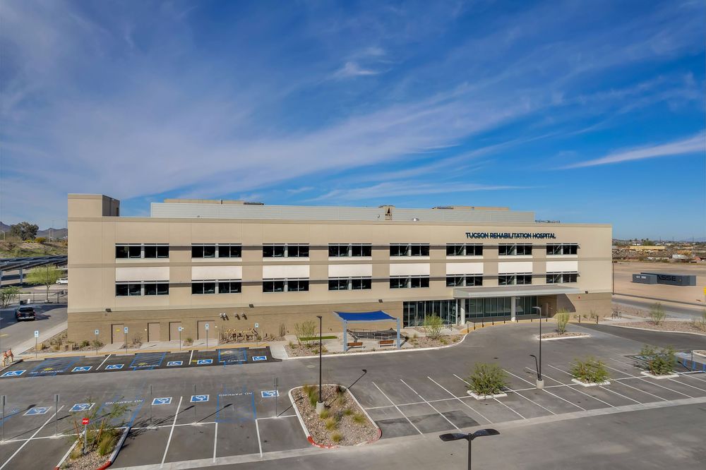 Panoramic view of a modern commercial building with tan exterior and large parking lot under bright blue desert skies.
