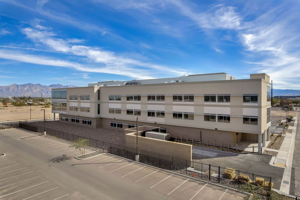 Modern three-story commercial office building with white exterior and large parking lot under bright blue desert sky.