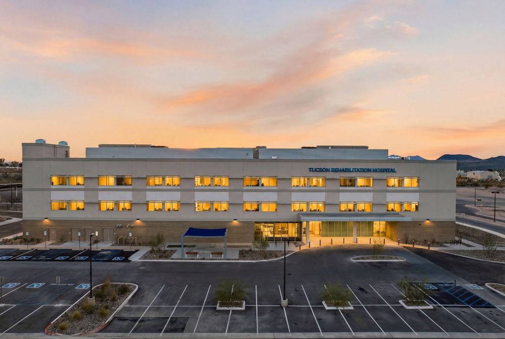Modern commercial building exterior with illuminated windows at sunset and empty parking lot in foreground.