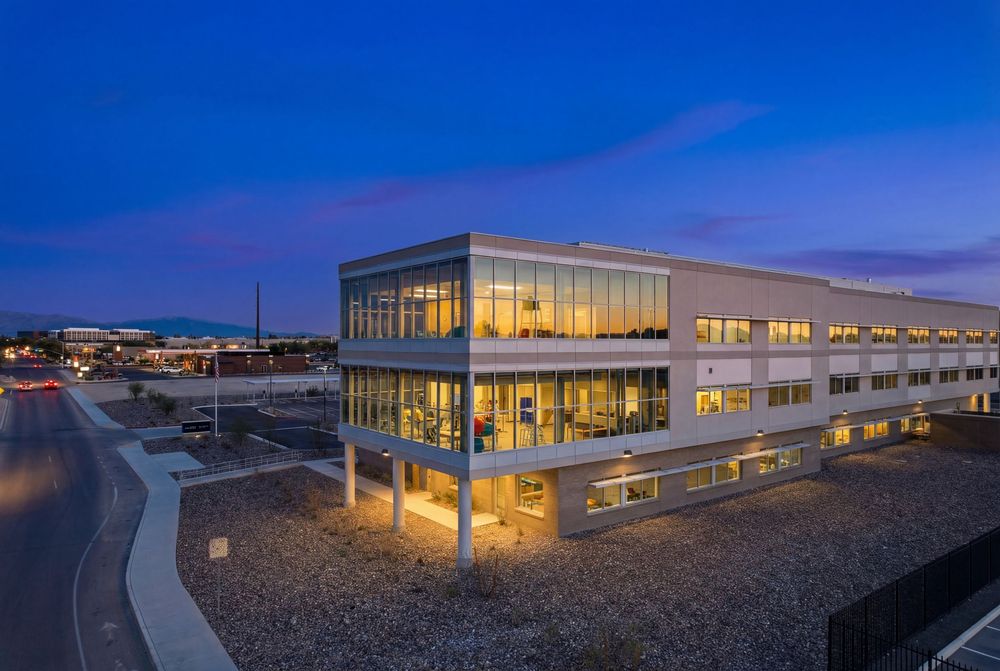 Modern glass building illuminated at dusk with expansive desert landscape and mountain views in the background.
