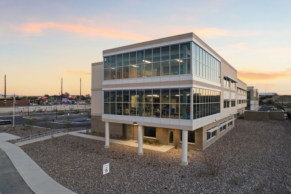 Modern office building exterior with glass windows and metal facade shown at sunset against a colorful sky.