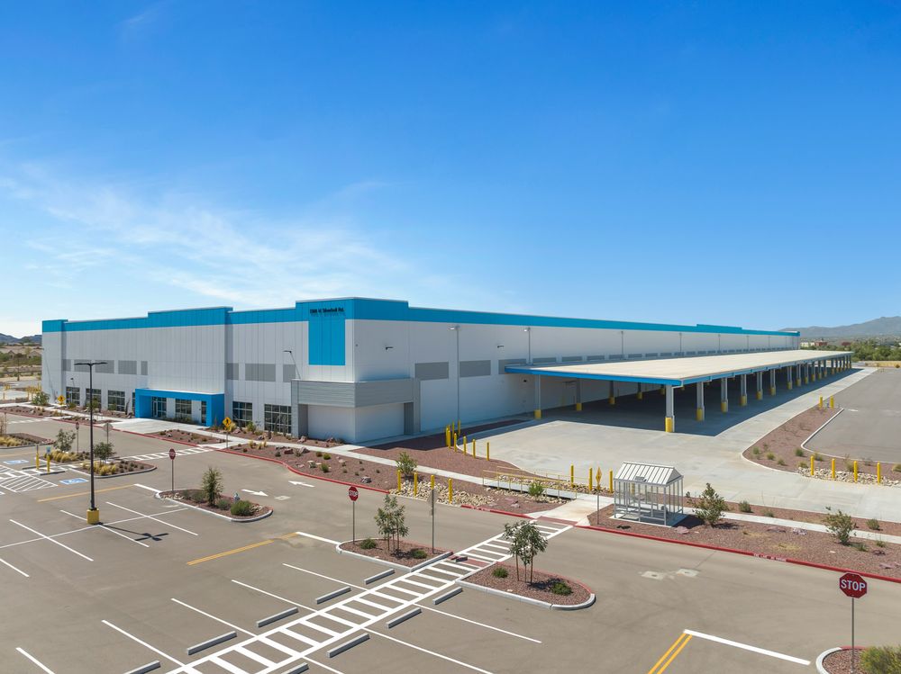 Modern industrial warehouse facility with loading docks and parking lot under bright blue sky in desert landscape.