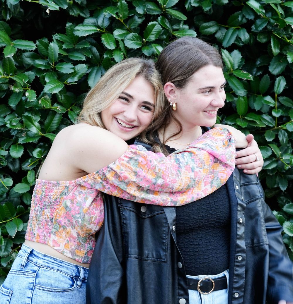 Two young women smiling and hugging outdoors in front of green leafy bushes, wearing casual stylish outfits.