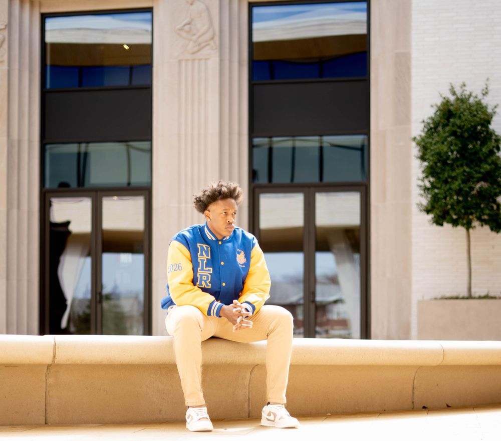 Young man in blue and yellow varsity jacket sits on stone ledge outside a building on a sunny day.