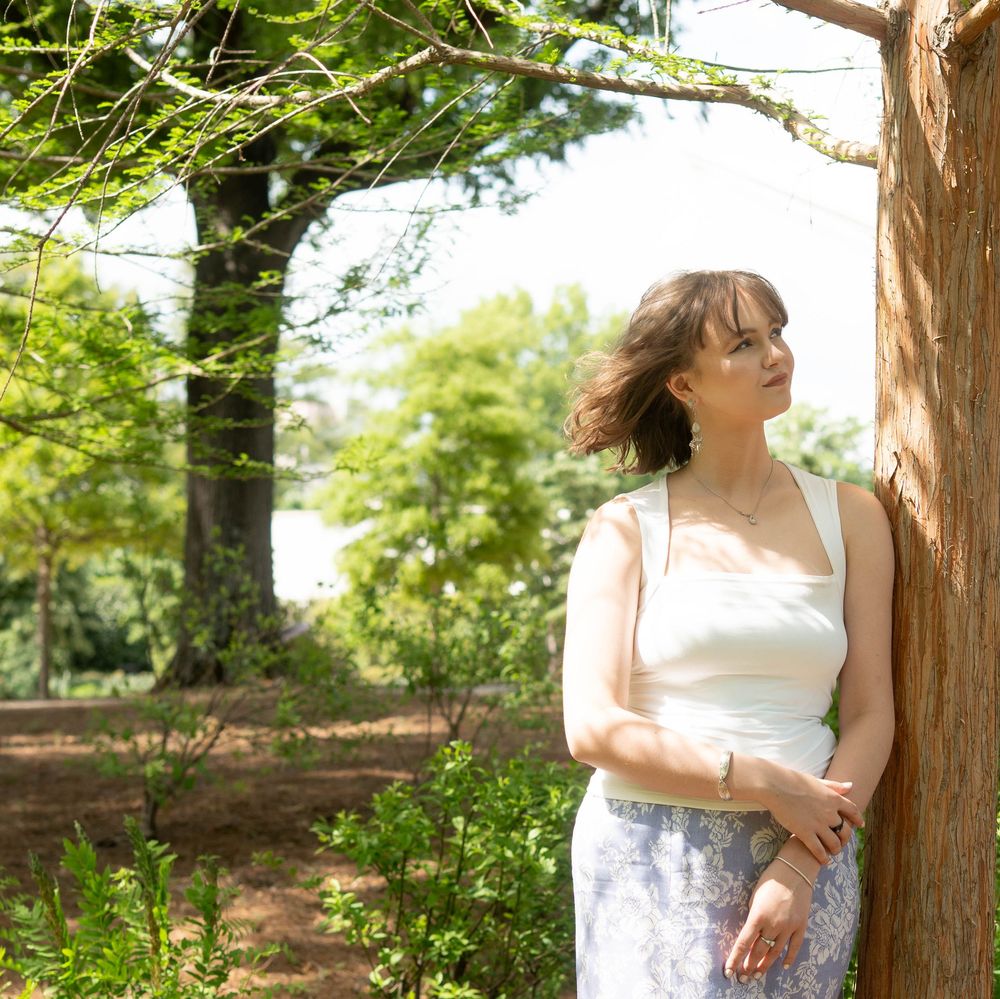 Young woman in white top and floral skirt leaning against tree in lush garden.