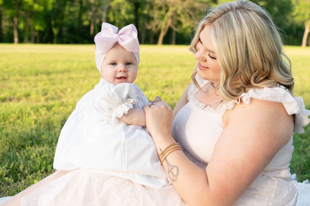 Blonde mother in pink dress holding smiling baby girl in white dress outdoors.