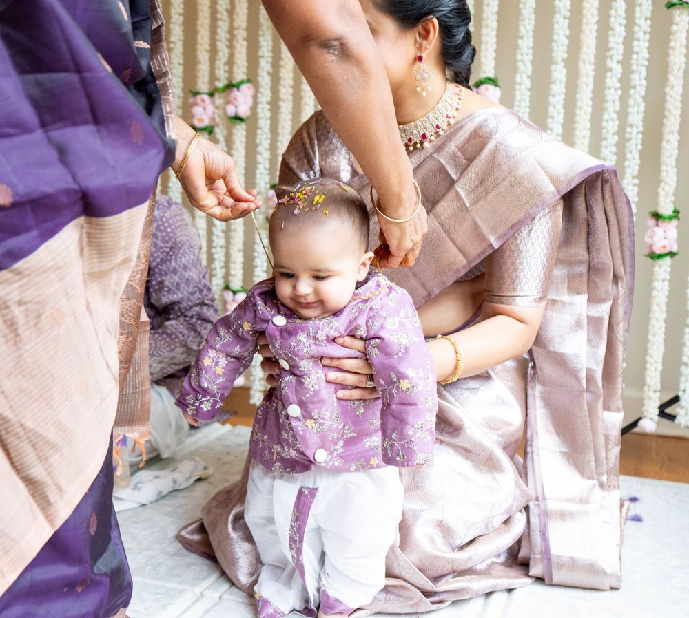 A smiling baby in purple floral outfit stands with help from a woman in a silk saree during an Indian ceremony.