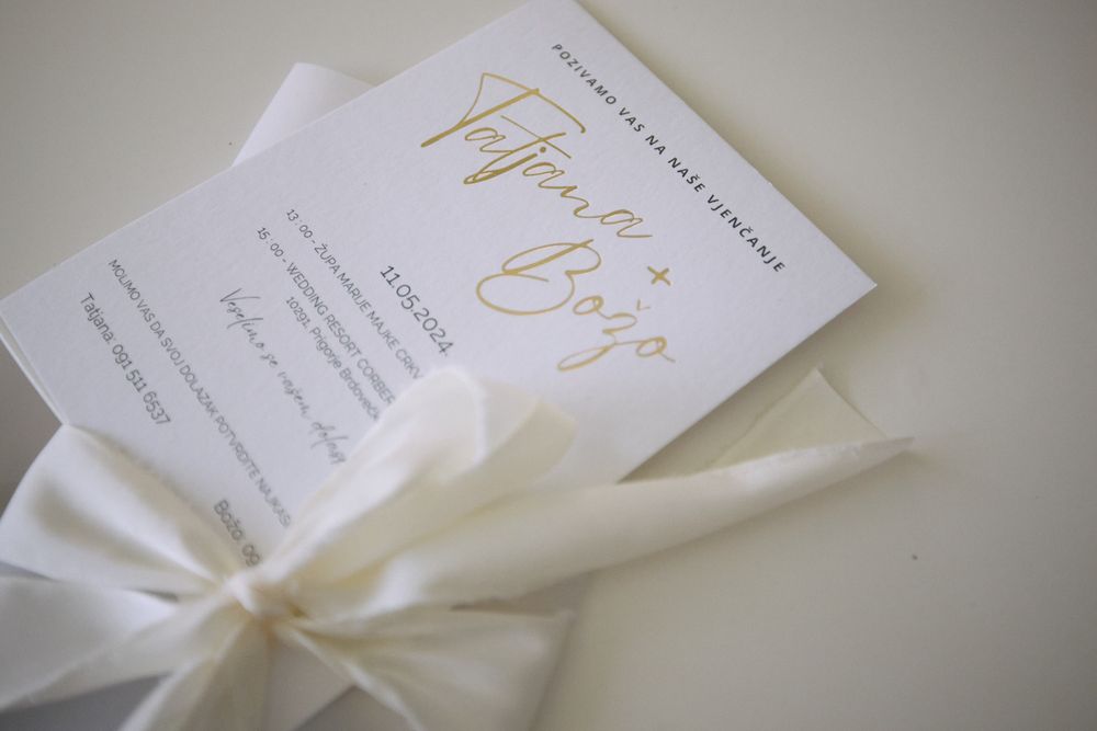 Elegant wedding invitation with gold script text and white silk ribbon on light gray background.