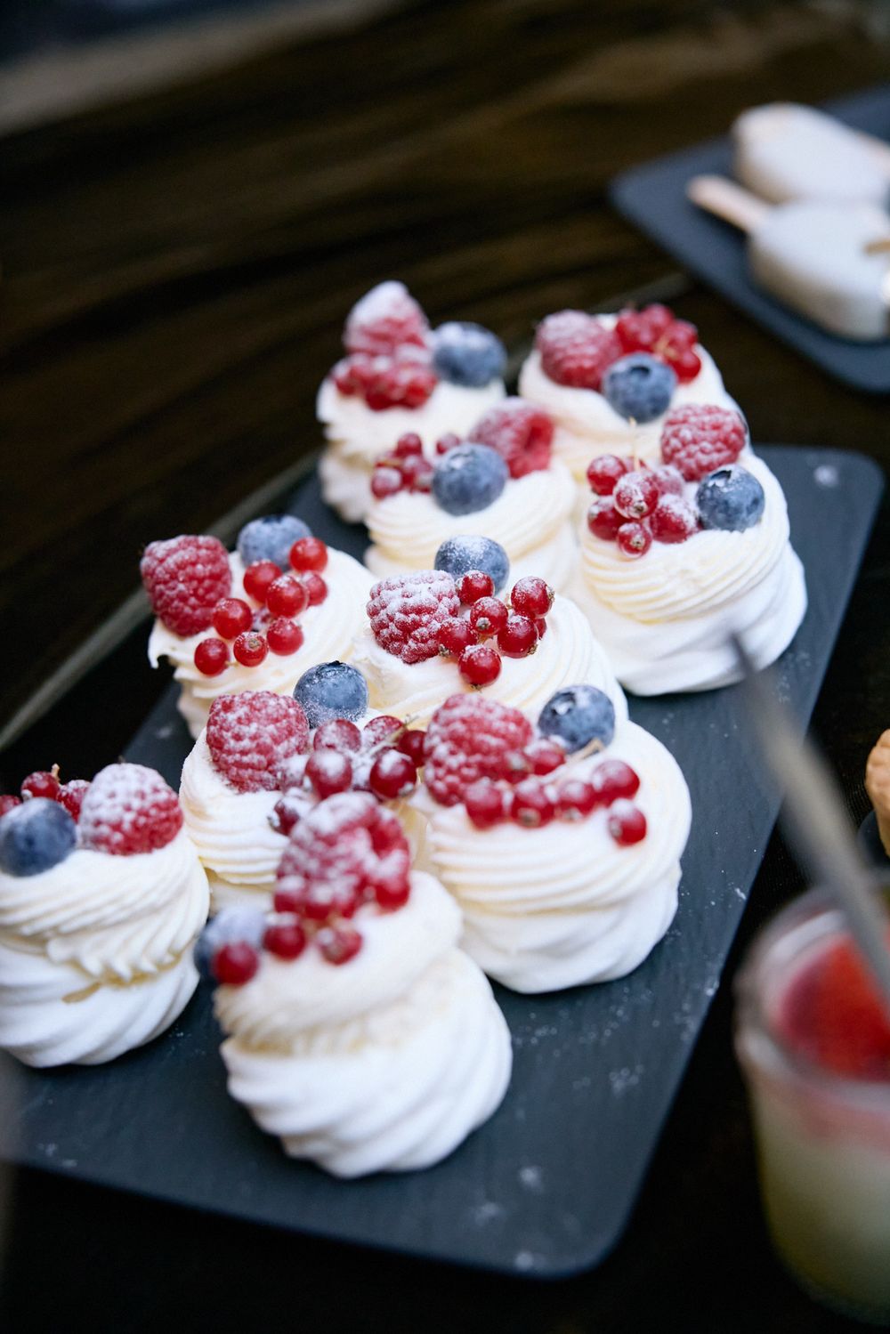 Mini pavlovas topped with fresh berries and powdered sugar on a dark serving platter.