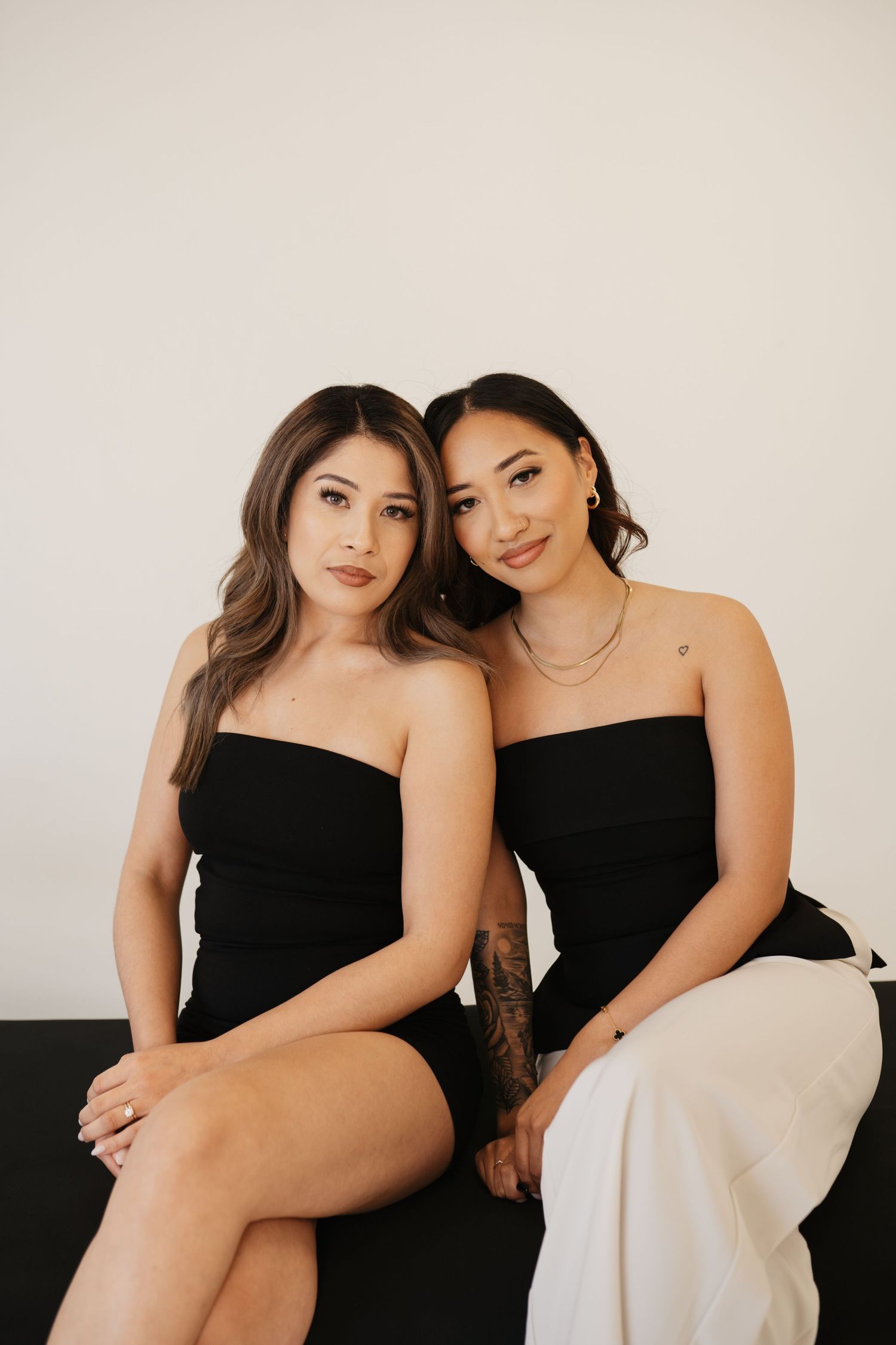 Two women in strapless dresses pose together on a dark couch against a light neutral background.