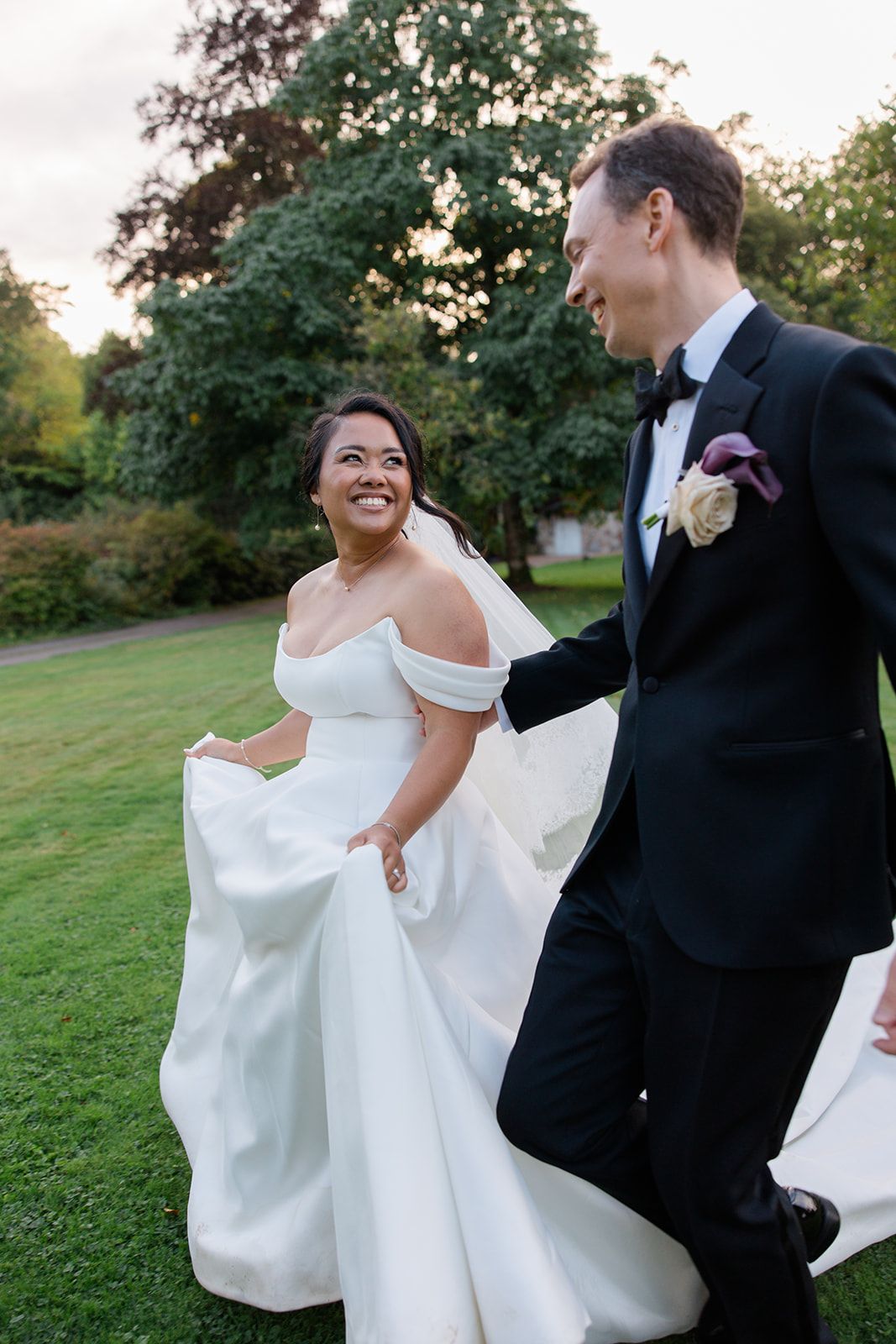 A couple in wedding attire share a joyful dance moment together on a grassy lawn at sunset.