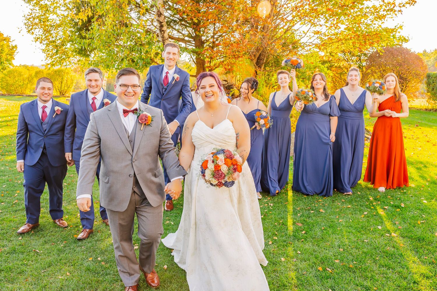 Wedding party stands together in autumn sunlight on grassy lawn during outdoor celebration.