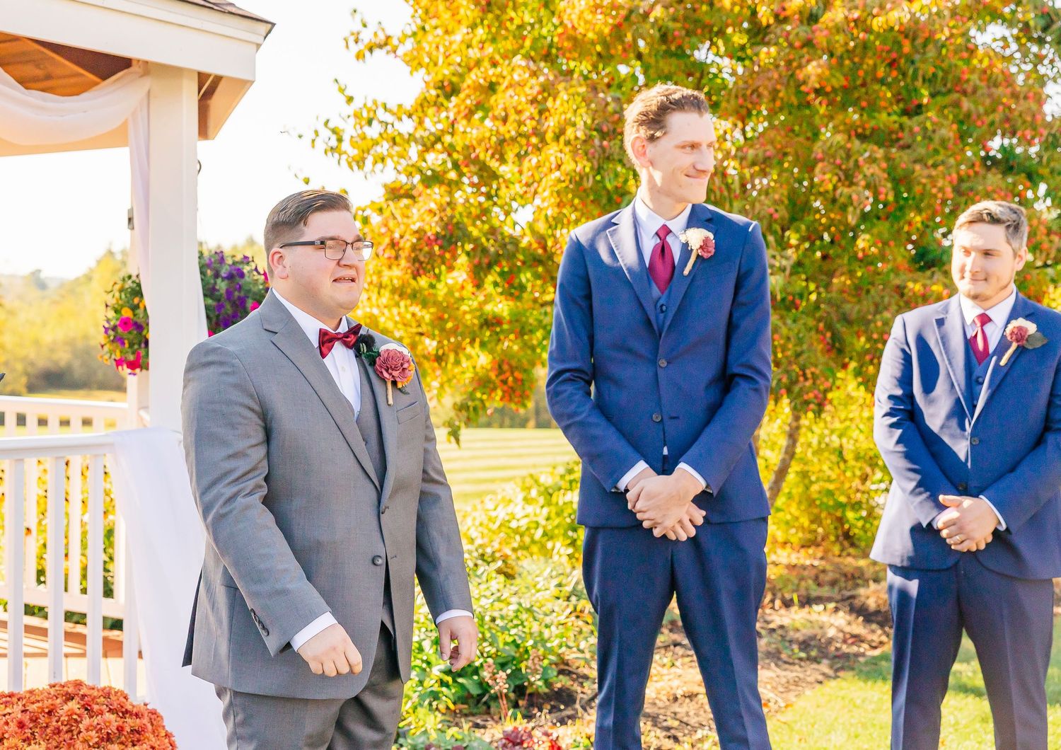 Wedding party members in blue suits and grey suit stand at outdoor ceremony with autumn foliage in background.