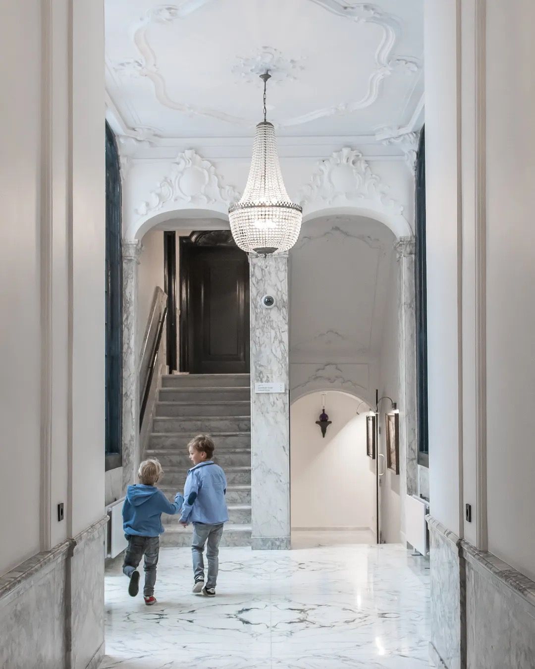 Bright marble hallway with a grand chandelier and two children walking toward an arched doorway.