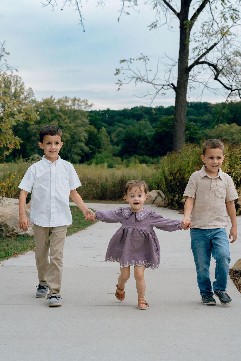 Three children walking hand in hand on a paved path surrounded by autumn trees for Family Photo Session in Akron, Ohio Metro Park