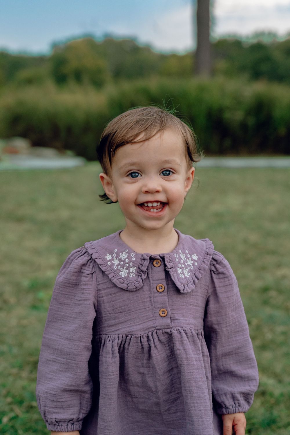 A toddler in a purple dress with a collared neckline smiles joyfully while playing outdoors in a grassy field in Akron, Ohio for Family Pics