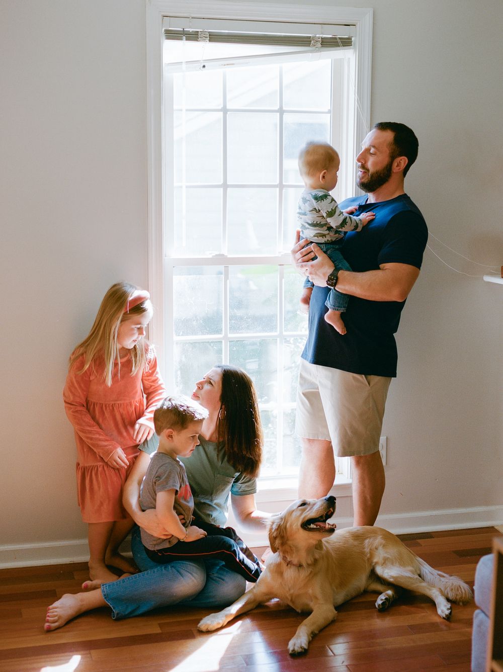 Family relaxes together by window with their golden retriever dog in a bright, sunlit room.