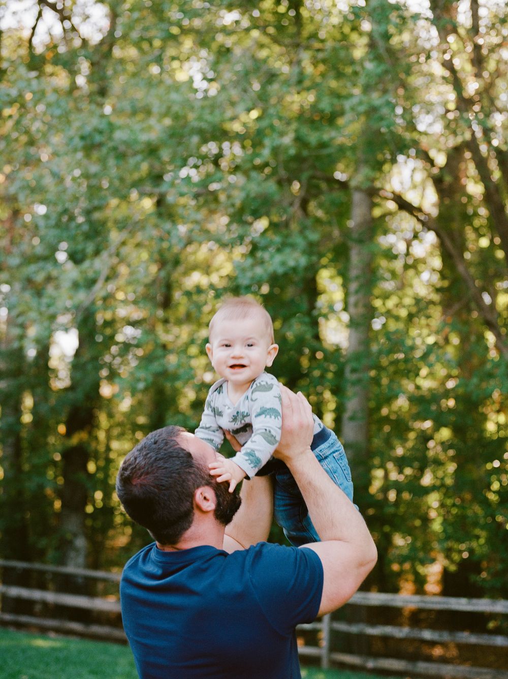 A joyful moment in a sunlit forest as a baby is lifted up high against a backdrop of green trees.