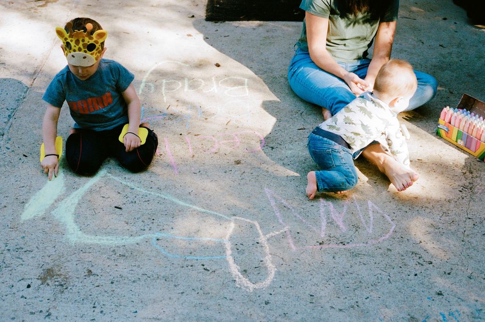 Children drawing with chalk on concrete pavement outdoors, one wearing a colorful mask.