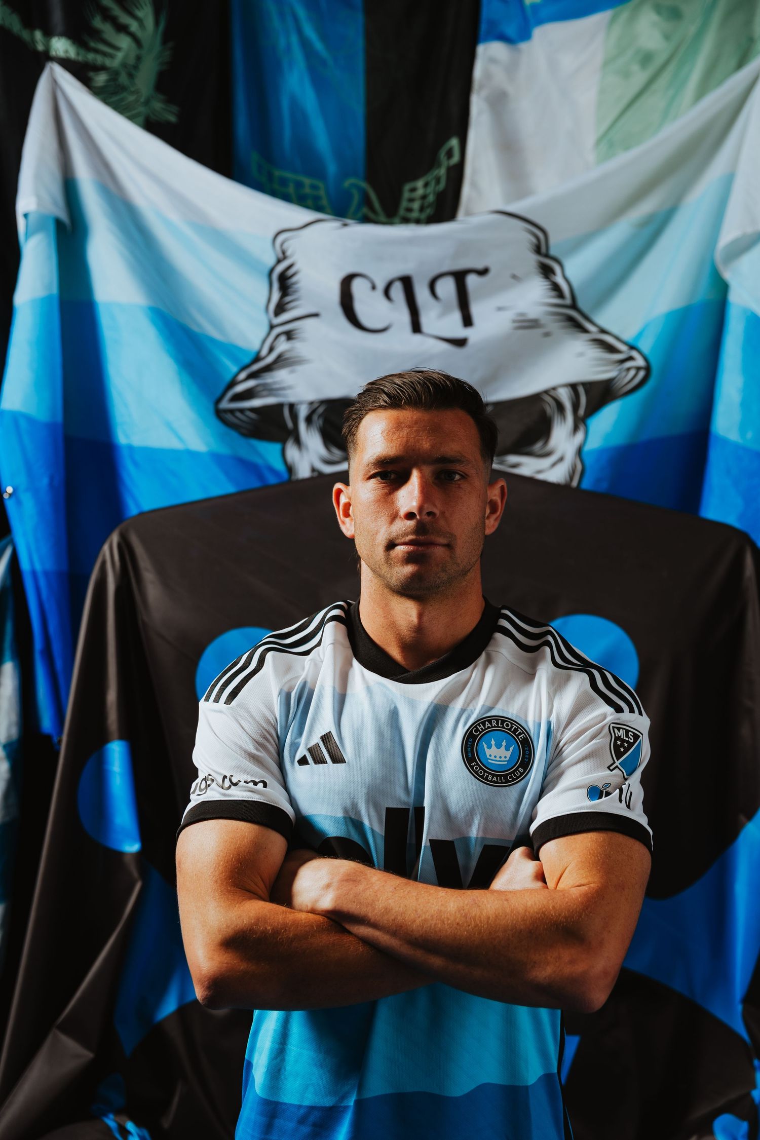 A Charlotte FC player in a white Adidas jersey poses with arms crossed before CIT supporter flags.