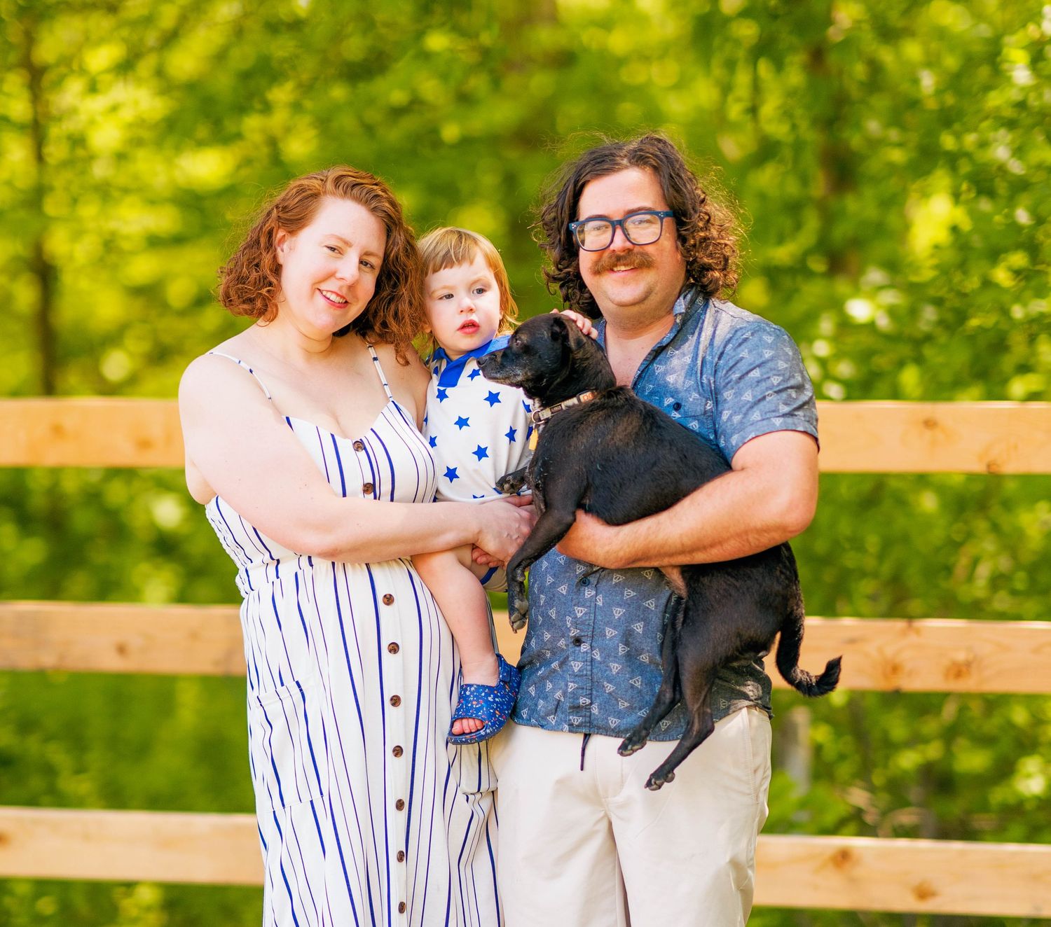 A family poses together with their black cat on a wooden fence during a sunny summer day in a green park setting.
