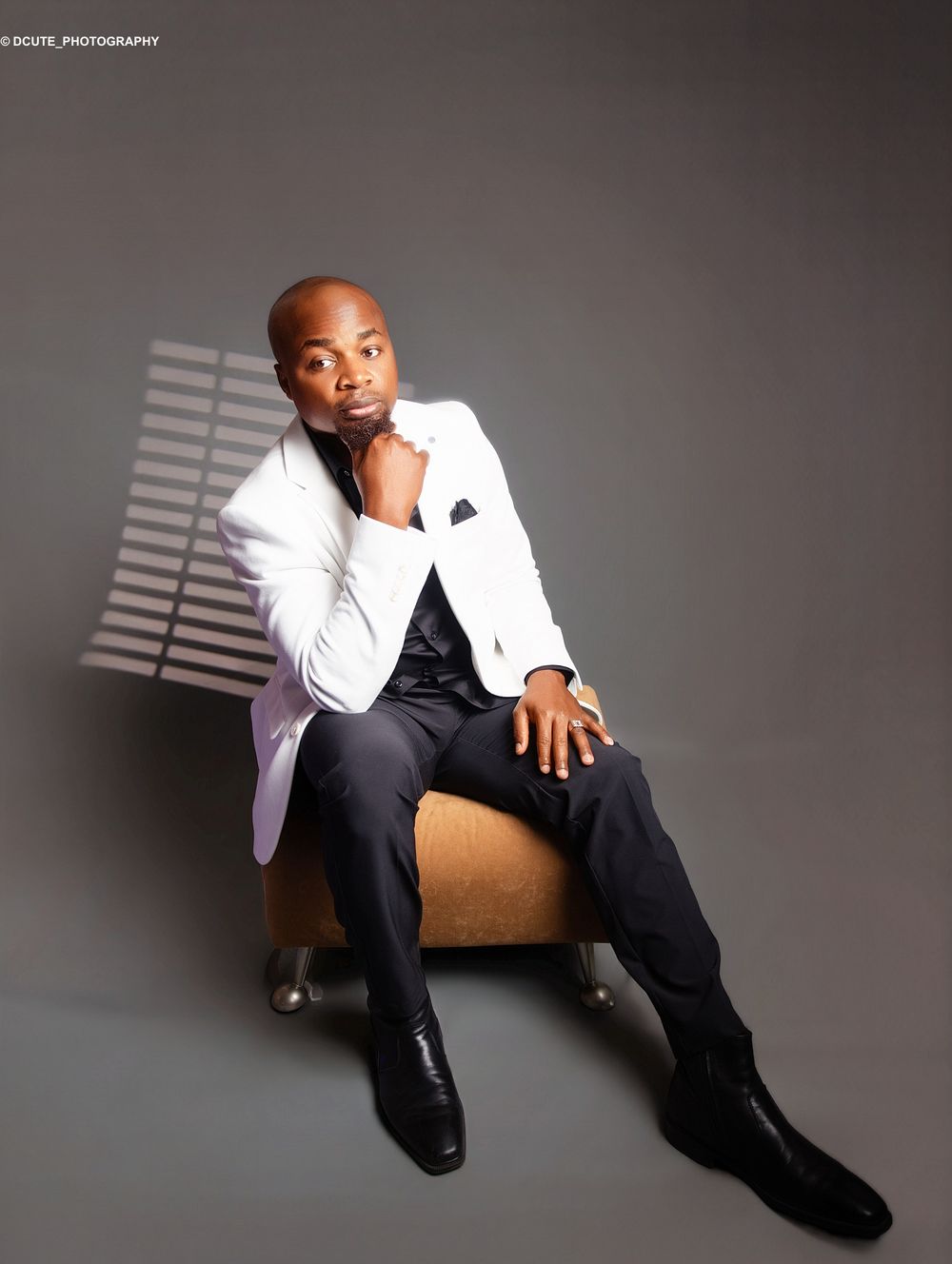 A stylish man in a white blazer and black outfit sits confidently on a chair in a professional photo shoot.