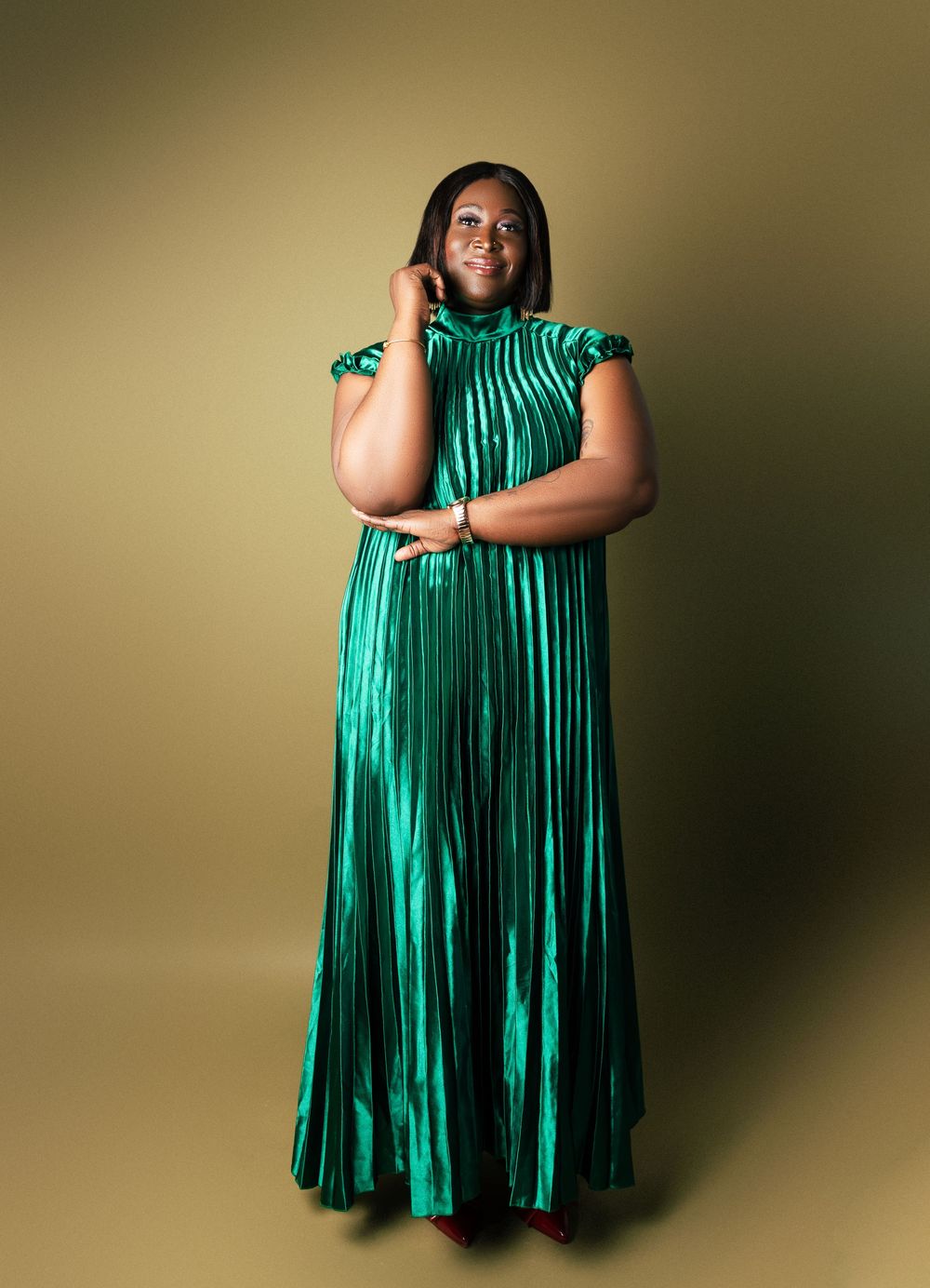Woman in elegant green pleated metallic maxi dress poses gracefully against a gold studio background.