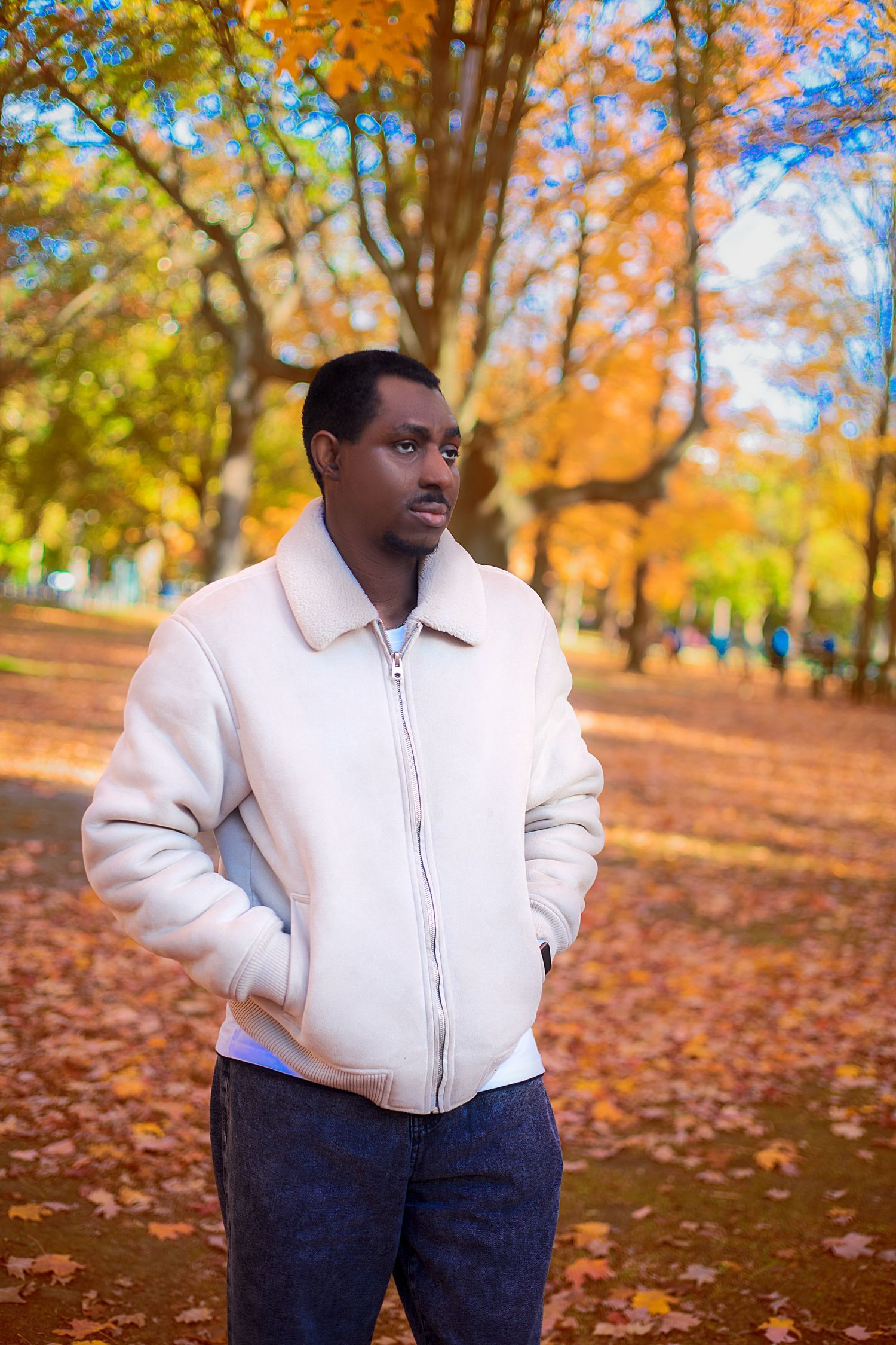 Black man in cream jacket stands in autumn park with colorful orange and yellow foliage.