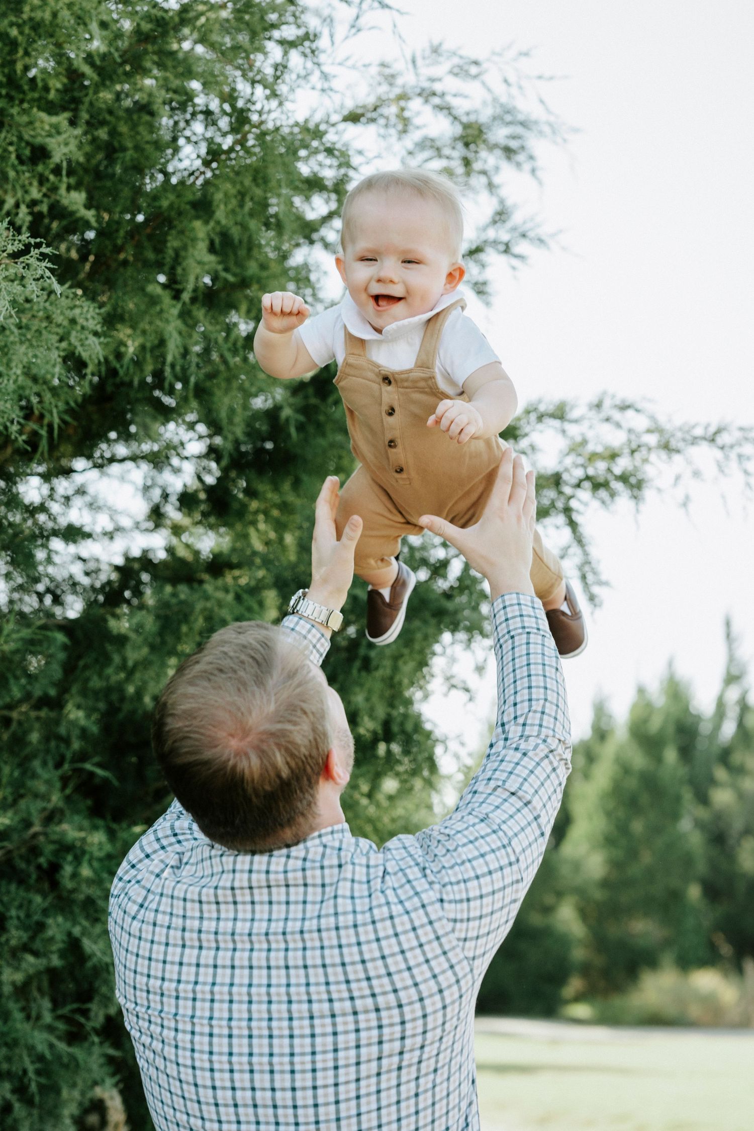 A playful moment captured outdoors as a baby wearing brown overalls is lifted joyfully into the air on a sunny day.
