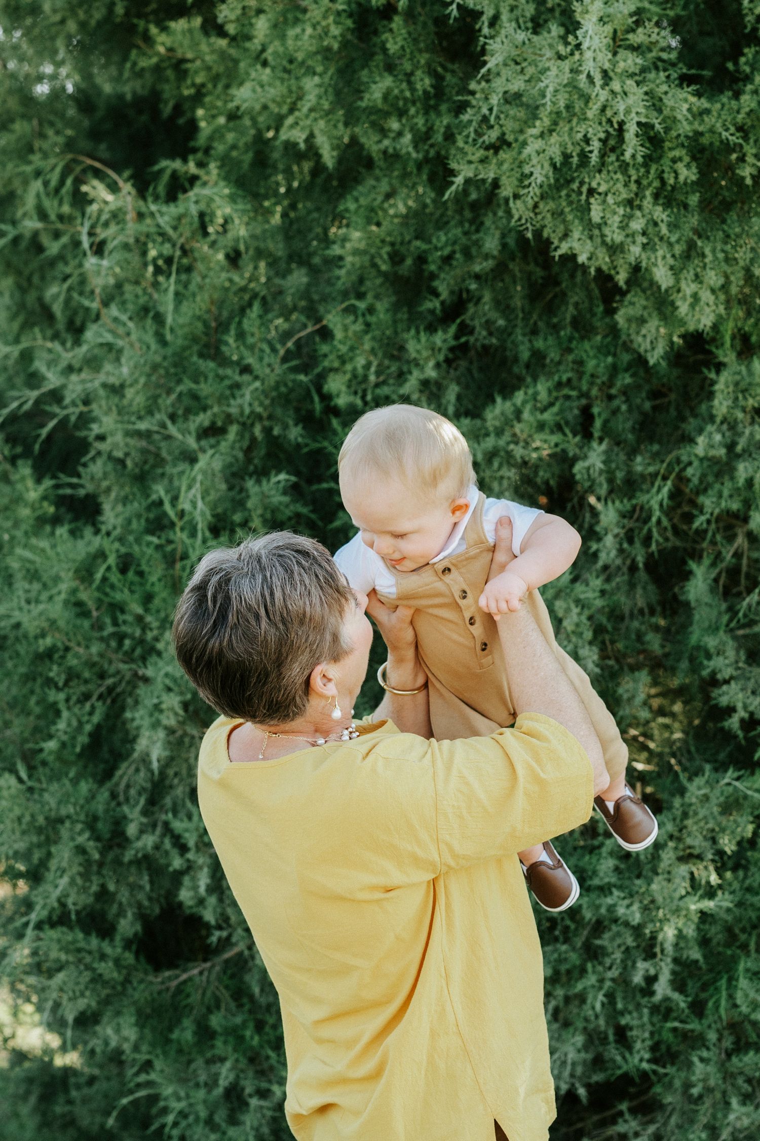 Parent in yellow shirt lifts and plays with small child against green foliage background in outdoor setting.