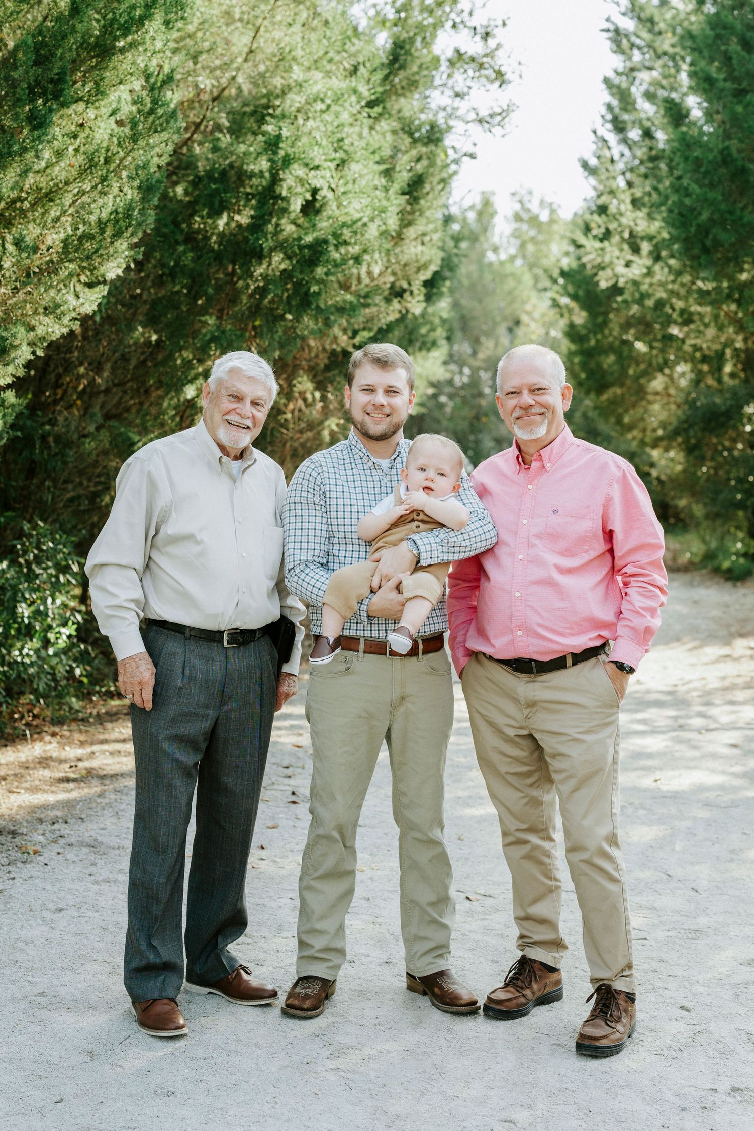 Four generations pose together on a sandy path surrounded by green trees in matching casual attire.