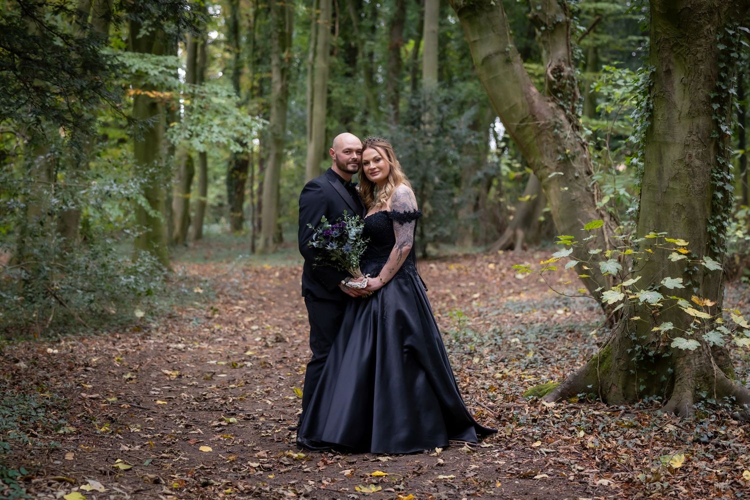 A couple in elegant black attire poses together in a lush autumn forest setting with fallen leaves on the ground.