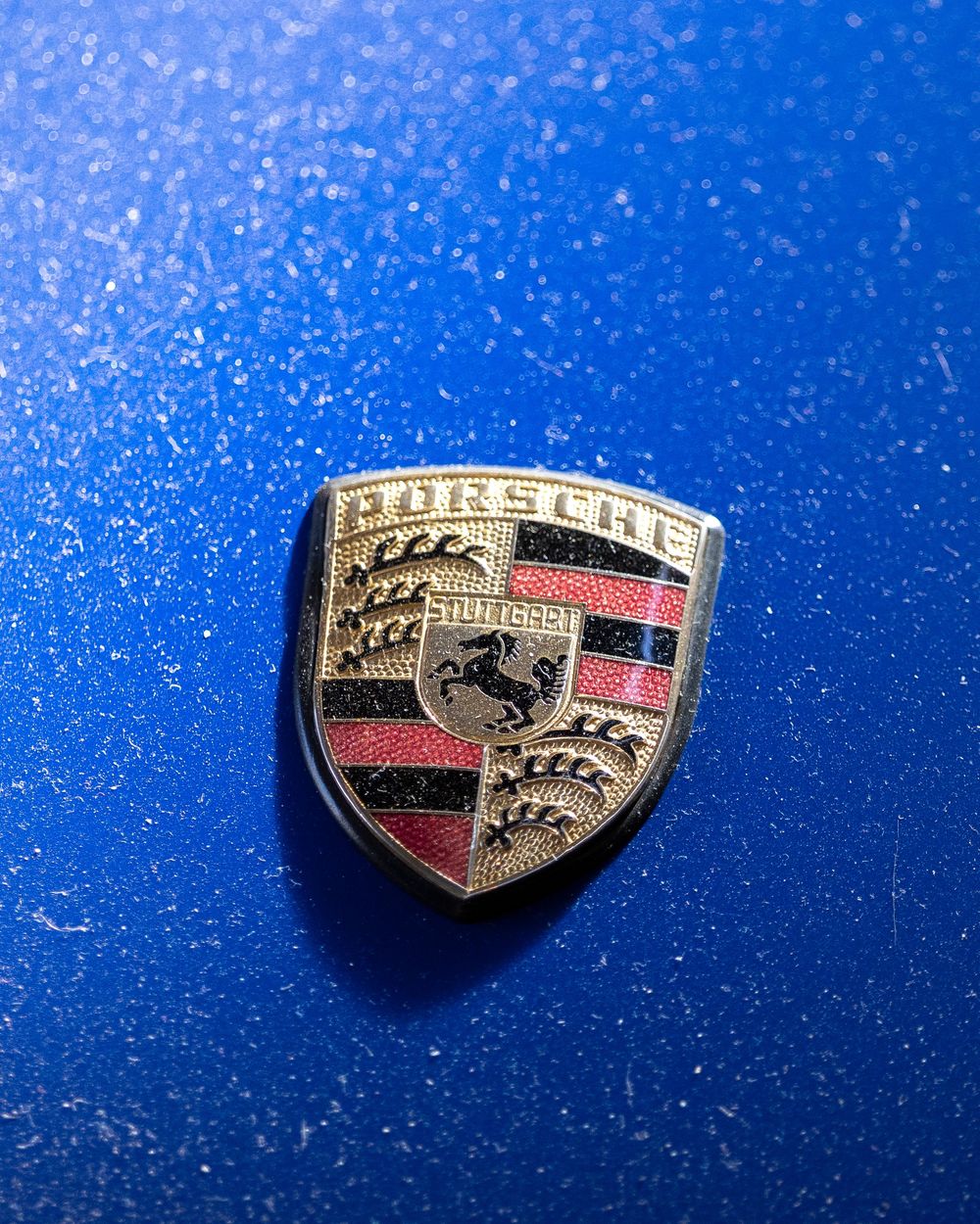 A series of four sequential photos showing a Porsche emblem badge on a blue metallic car surface fading into darkness.