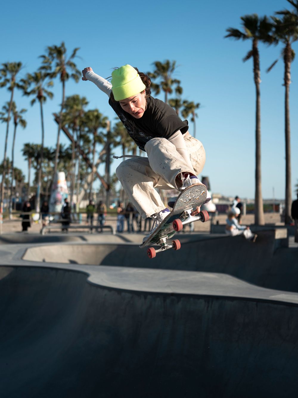 A skateboarder in a green beanie performs an aerial trick at a beachside skate park with palm trees in the background.