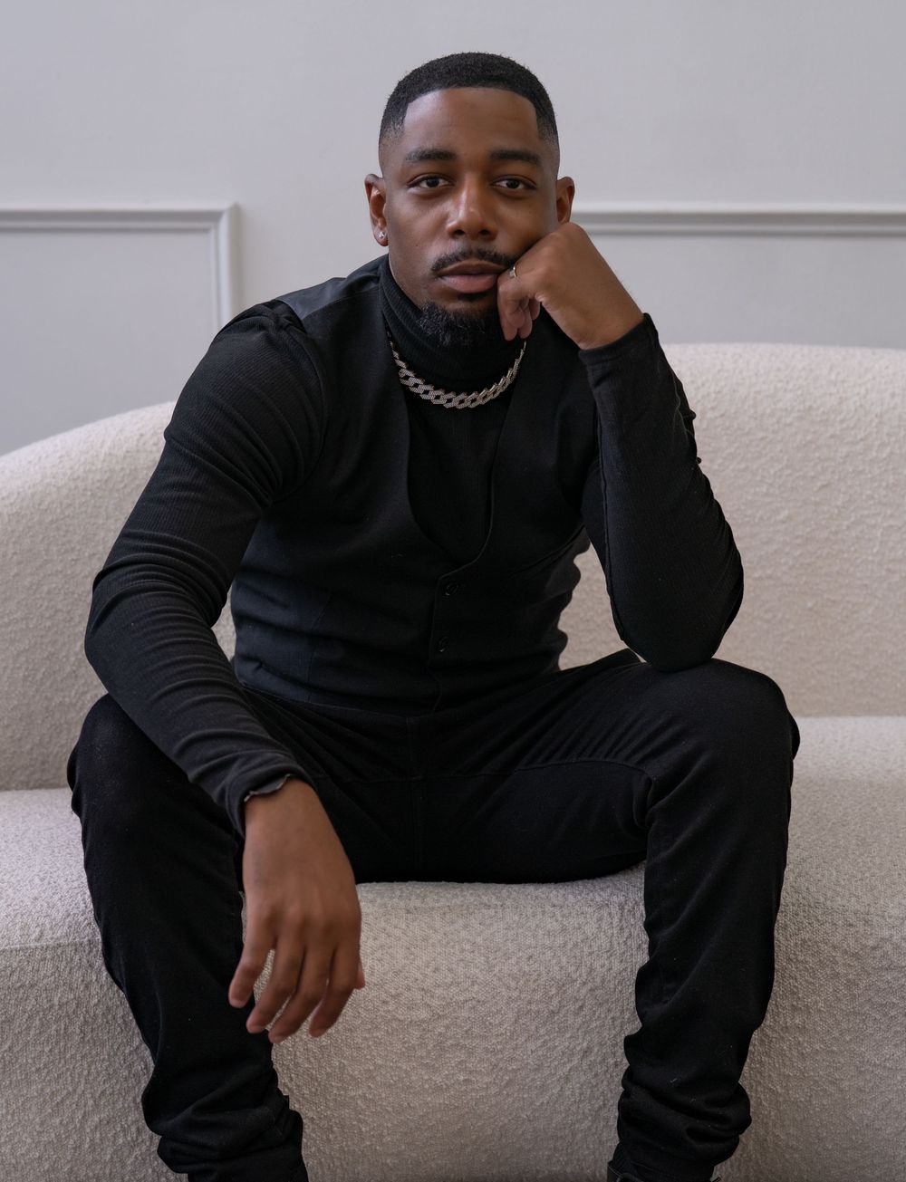 A person wearing all black sits on a white couch in a contemplative pose against a light gray wall.
